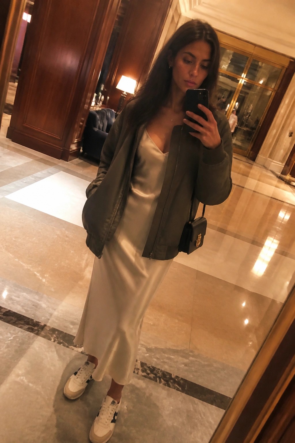 Woman in green bomber jacket over white slip dress taking mirror selfie in lobby