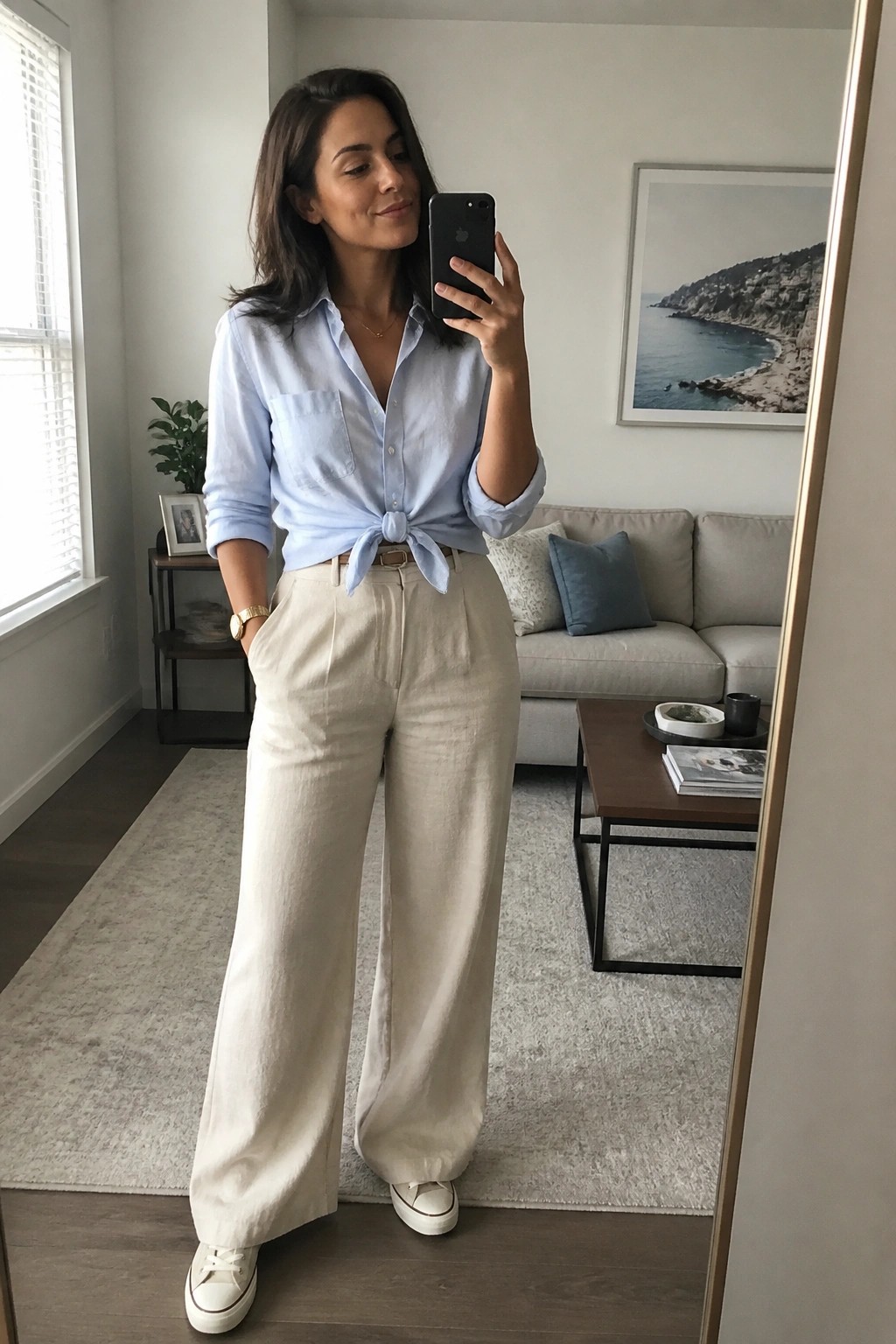 Woman in light blue tied shirt and cream wide-leg pants taking mirror selfie