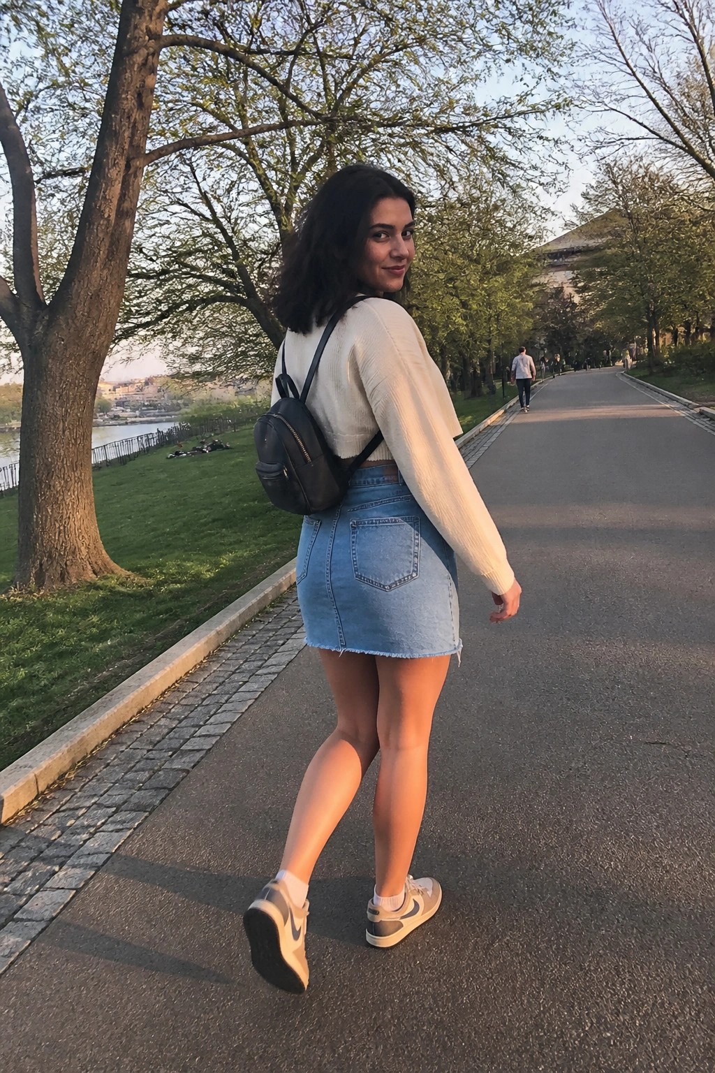 Woman in denim skirt and white sweater with backpack on park path, glancing back