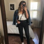 Woman in mirror selfie wearing blue denim jacket over cream satin camisole, black straight leg jeans, white sneakers, and black crossbody bag.