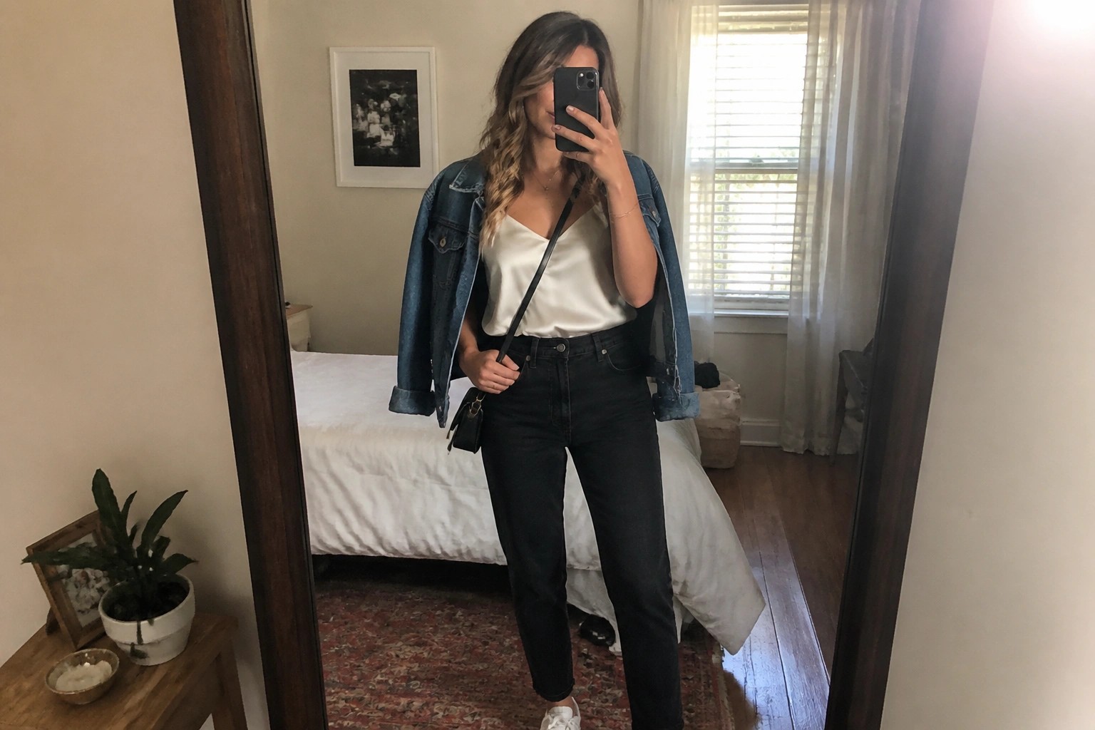 Woman in mirror selfie wearing blue denim jacket over cream satin camisole, black straight leg jeans, white sneakers, and black crossbody bag.