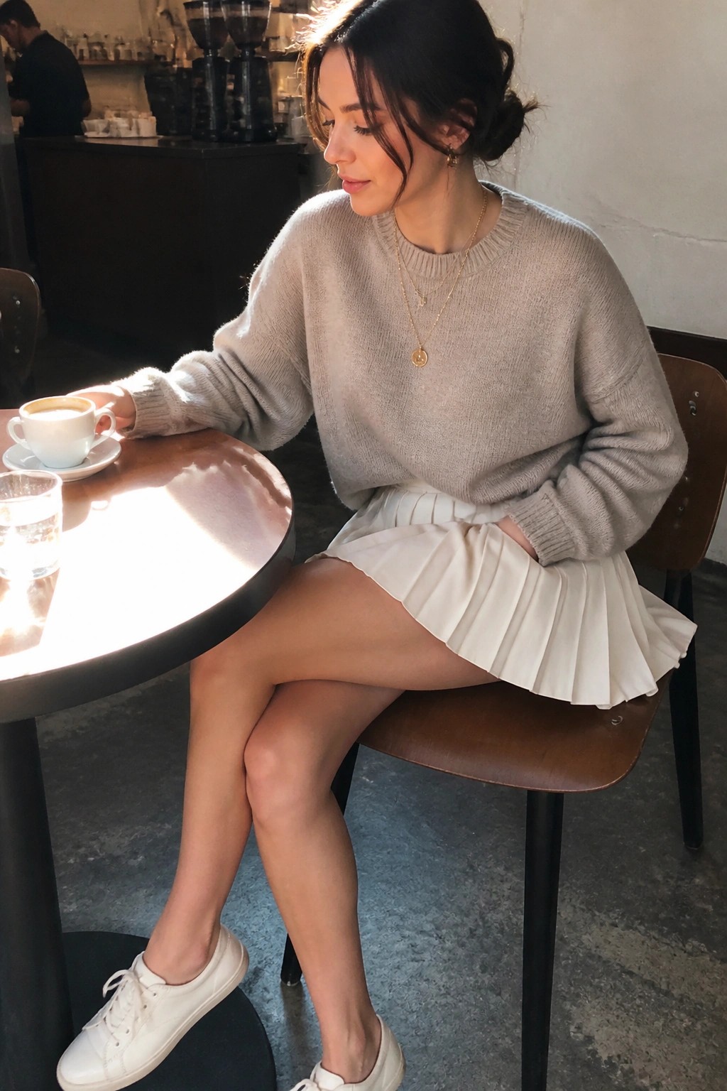 Woman in beige sweater and white pleated skirt at sunny cafe table with coffee