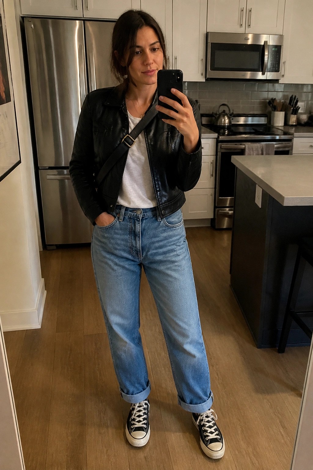 Woman in black leather jacket over white t-shirt with cuffed blue jeans and black low-top Converse sneakers standing in a kitchen.