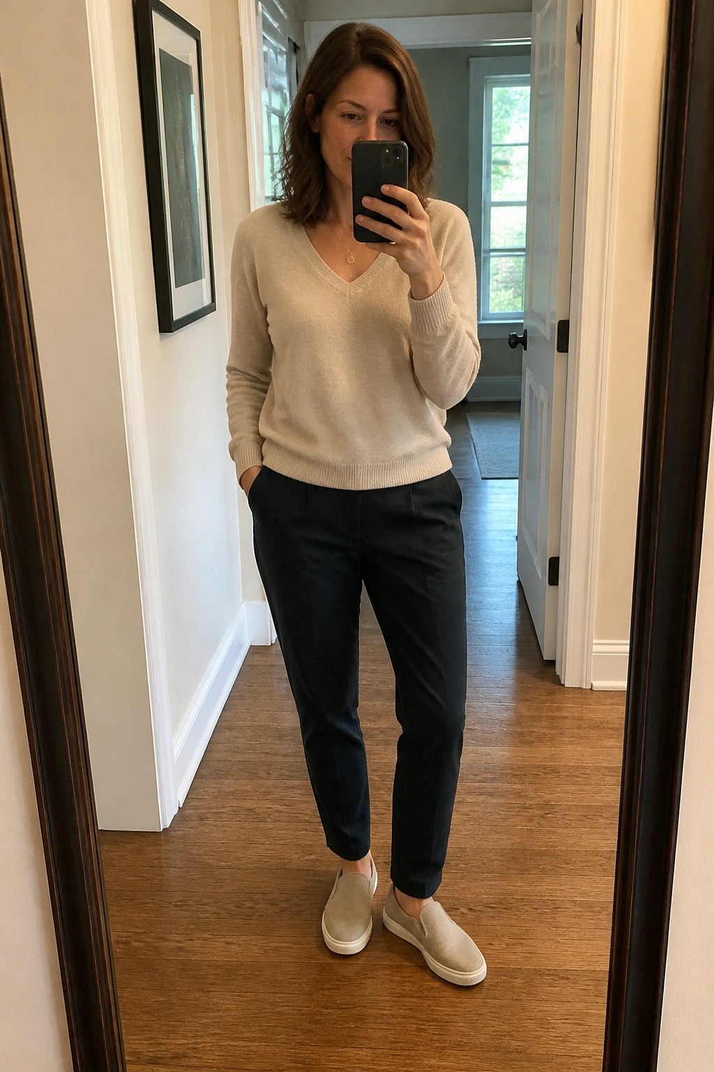 Woman in beige sweater and navy pants taking hallway mirror selfie.