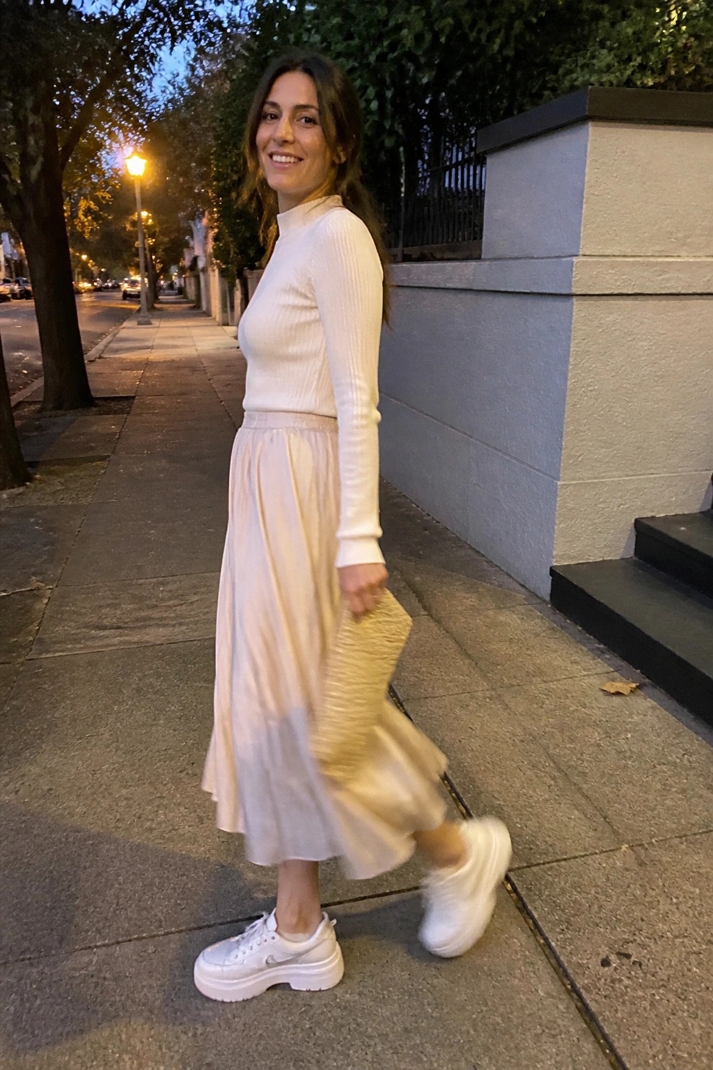 Smiling woman in white sweater and flowing skirt on city sidewalk at dusk