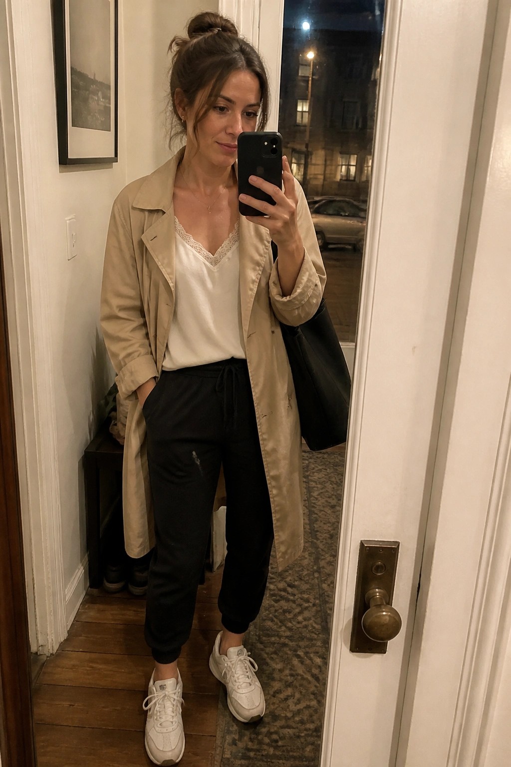 Woman wearing an open beige trench coat over a white lace camisole with black jogger pants and white sneakers while holding a phone for a mirror selfie.
