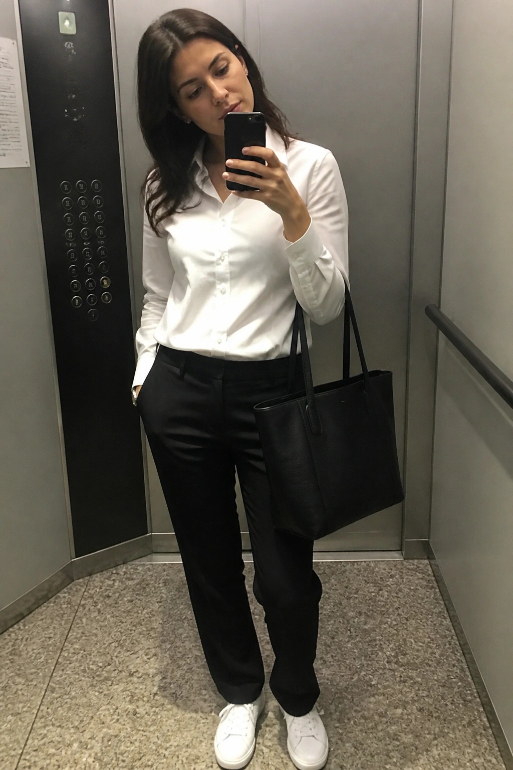 Woman wearing a white button-down shirt tucked into black trousers with white sneakers and carrying a black tote bag while taking a mirror selfie.
