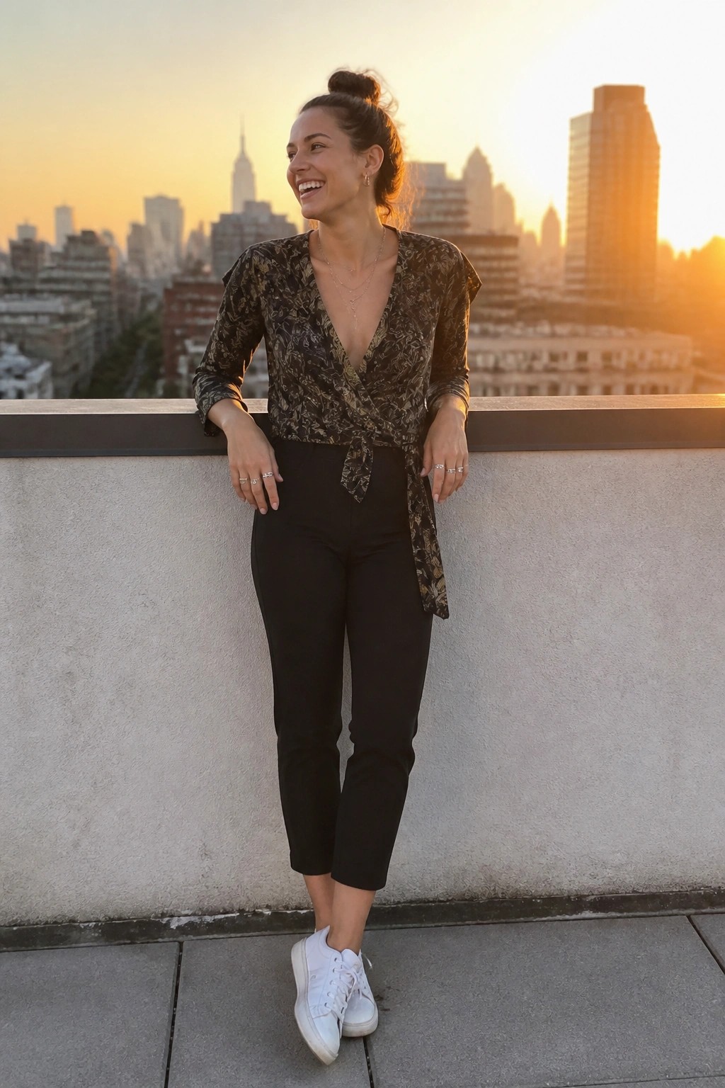Woman in patterned wrap top and black pants smiling on rooftop at sunset