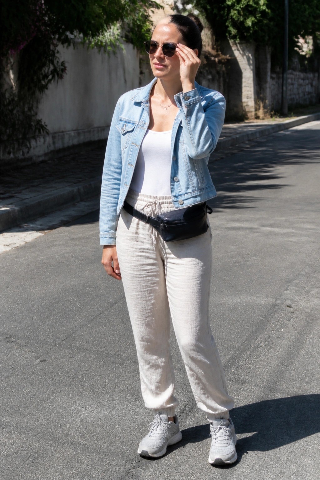 Woman wearing light blue denim jacket over white tank top with beige linen pants, white sneakers, and black belt bag.