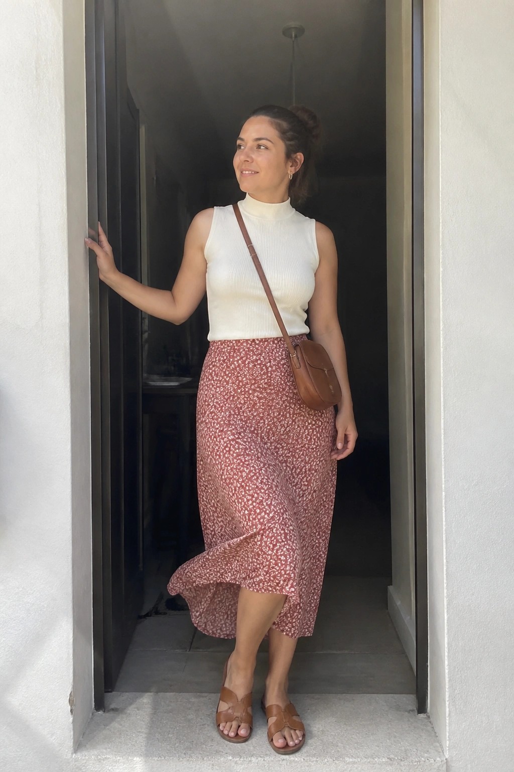 Woman in white sleeveless top and floral midi skirt in doorway with brown bag