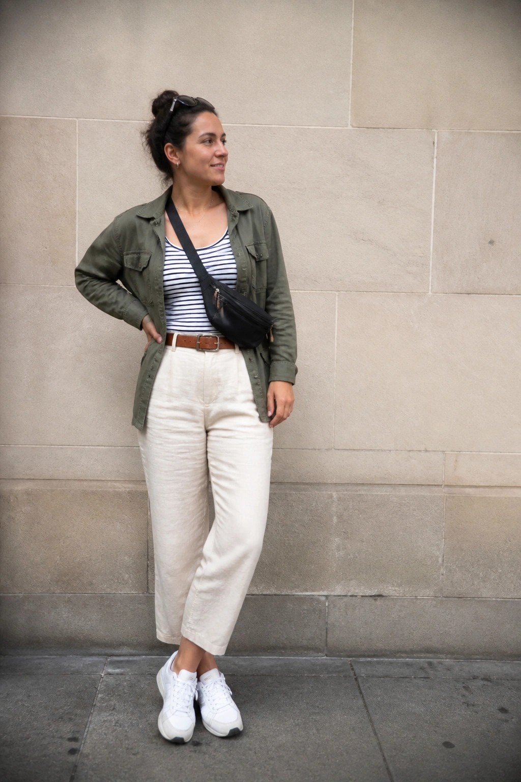 Woman in olive utility jacket over striped tank top, cream wide leg pants with brown belt, black crossbody bag, and white sneakers against stone wall.