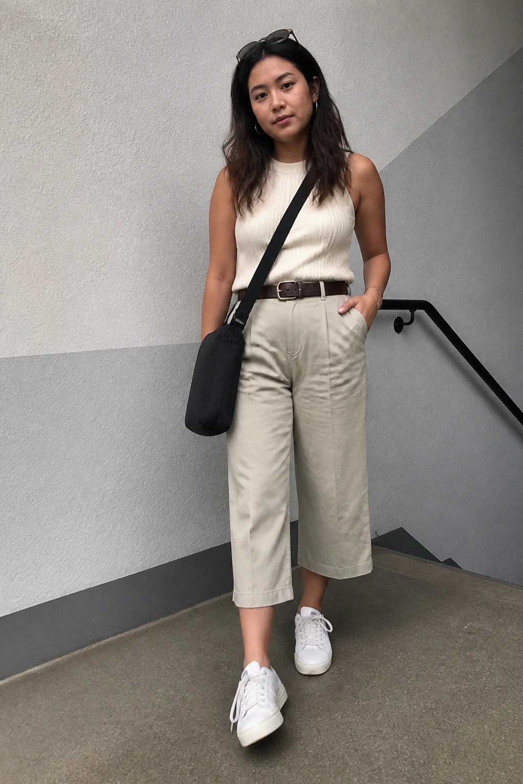 Woman in cream sleeveless top, beige cropped pants, brown belt, white sneakers, black bag