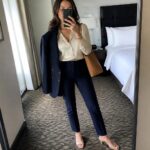 Woman wearing a navy blue tailored suit with an open blazer over a white button-up shirt, paired with navy trousers, nude block heel pumps, and a brown leather tote bag.