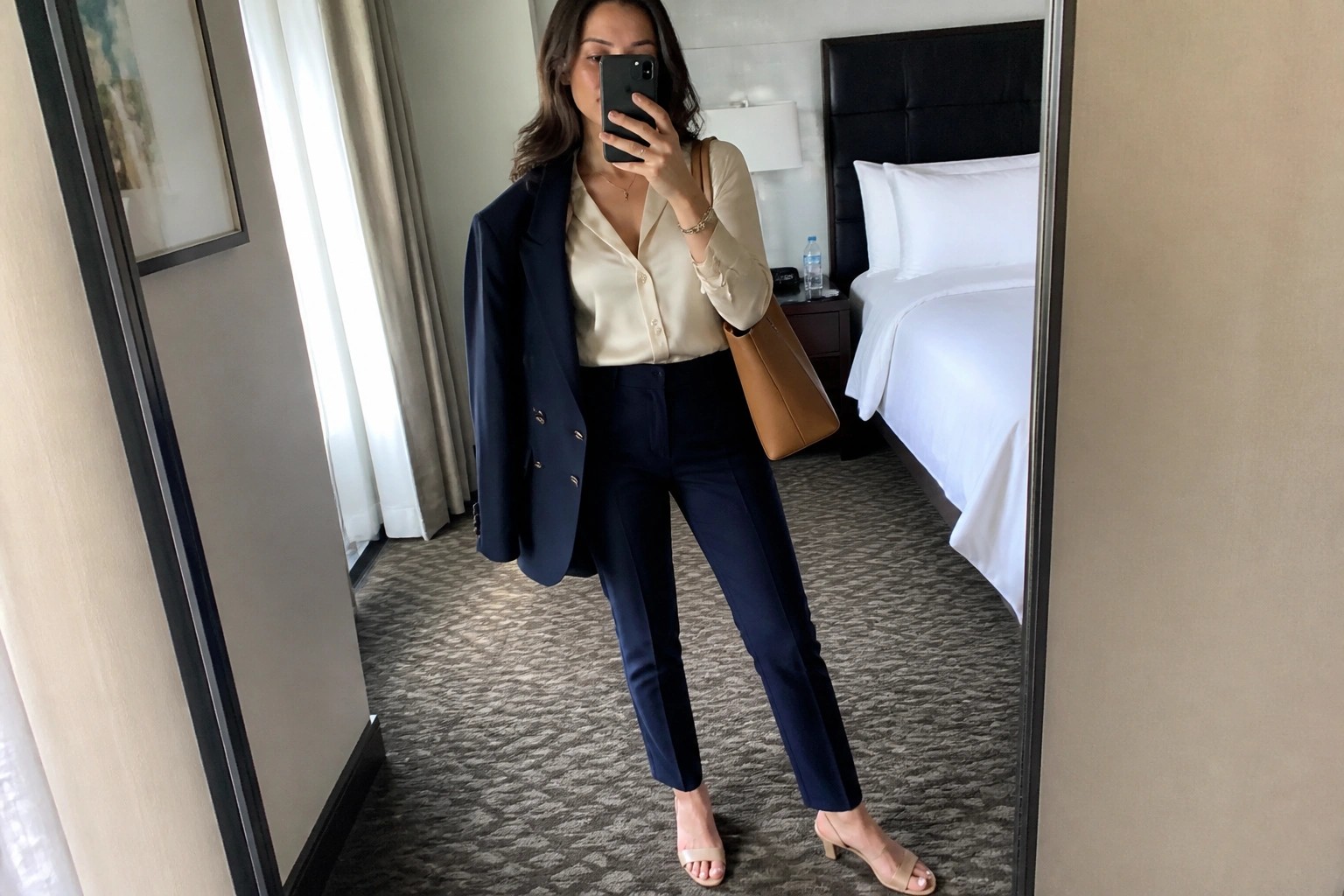 Woman wearing a navy blue tailored suit with an open blazer over a white button-up shirt, paired with navy trousers, nude block heel pumps, and a brown leather tote bag.