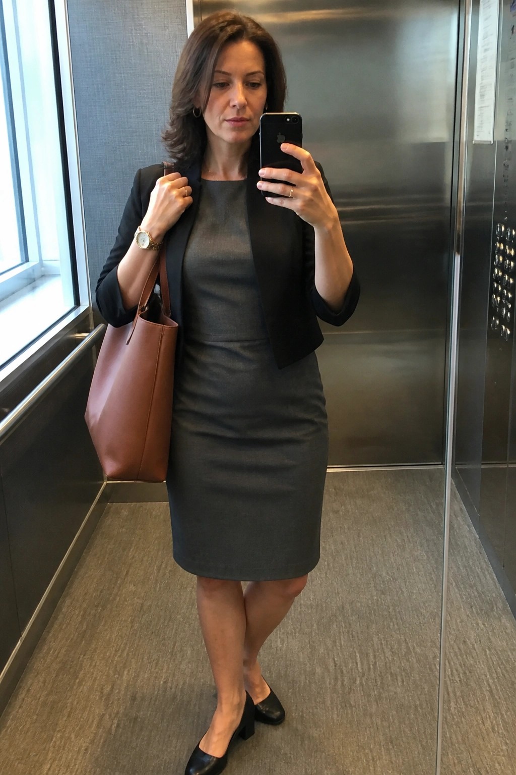 Woman in gray dress and black blazer taking selfie in elevator with brown bag