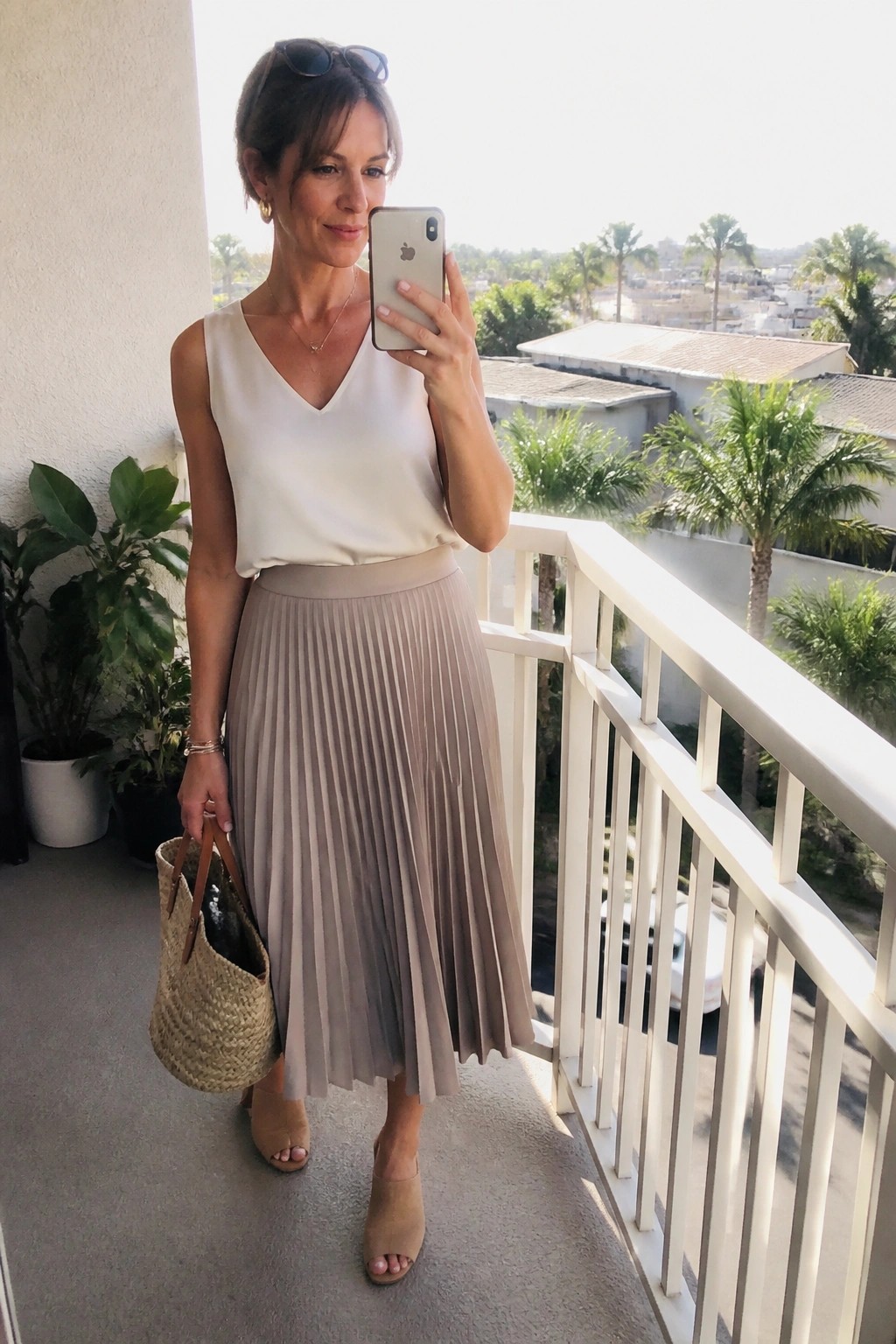Woman in white top and beige pleated skirt taking selfie on balcony with straw bag