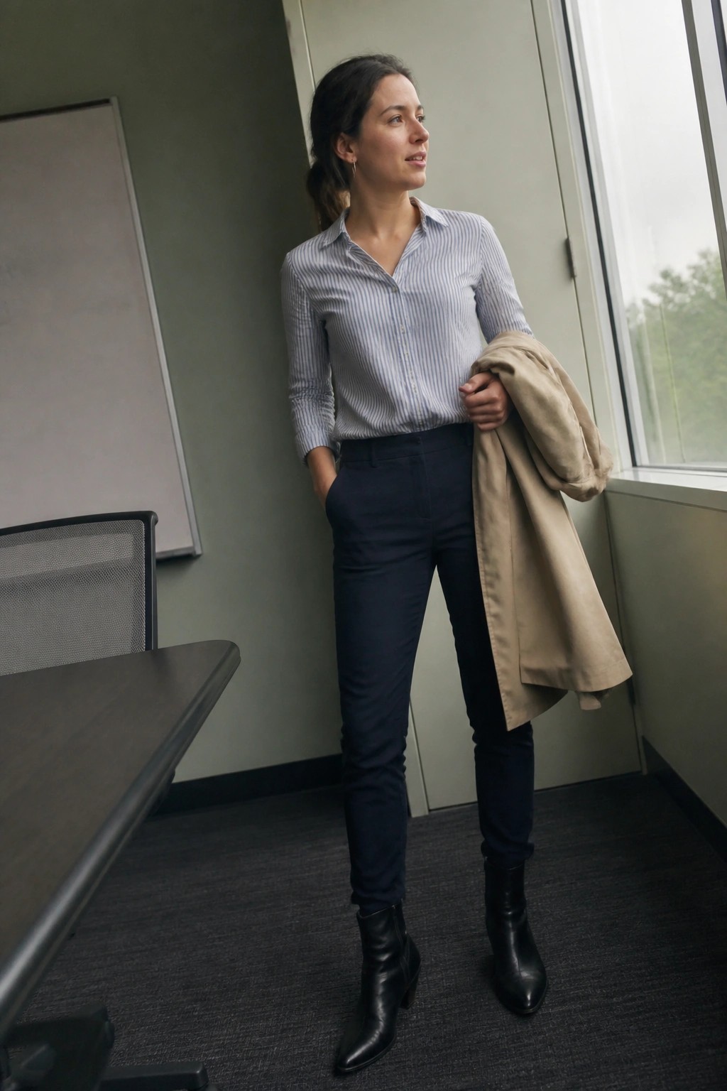 Woman in a blue and white striped button down shirt tucked into slim navy pants wearing black ankle boots and holding a beige coat over one arm in an office setting.