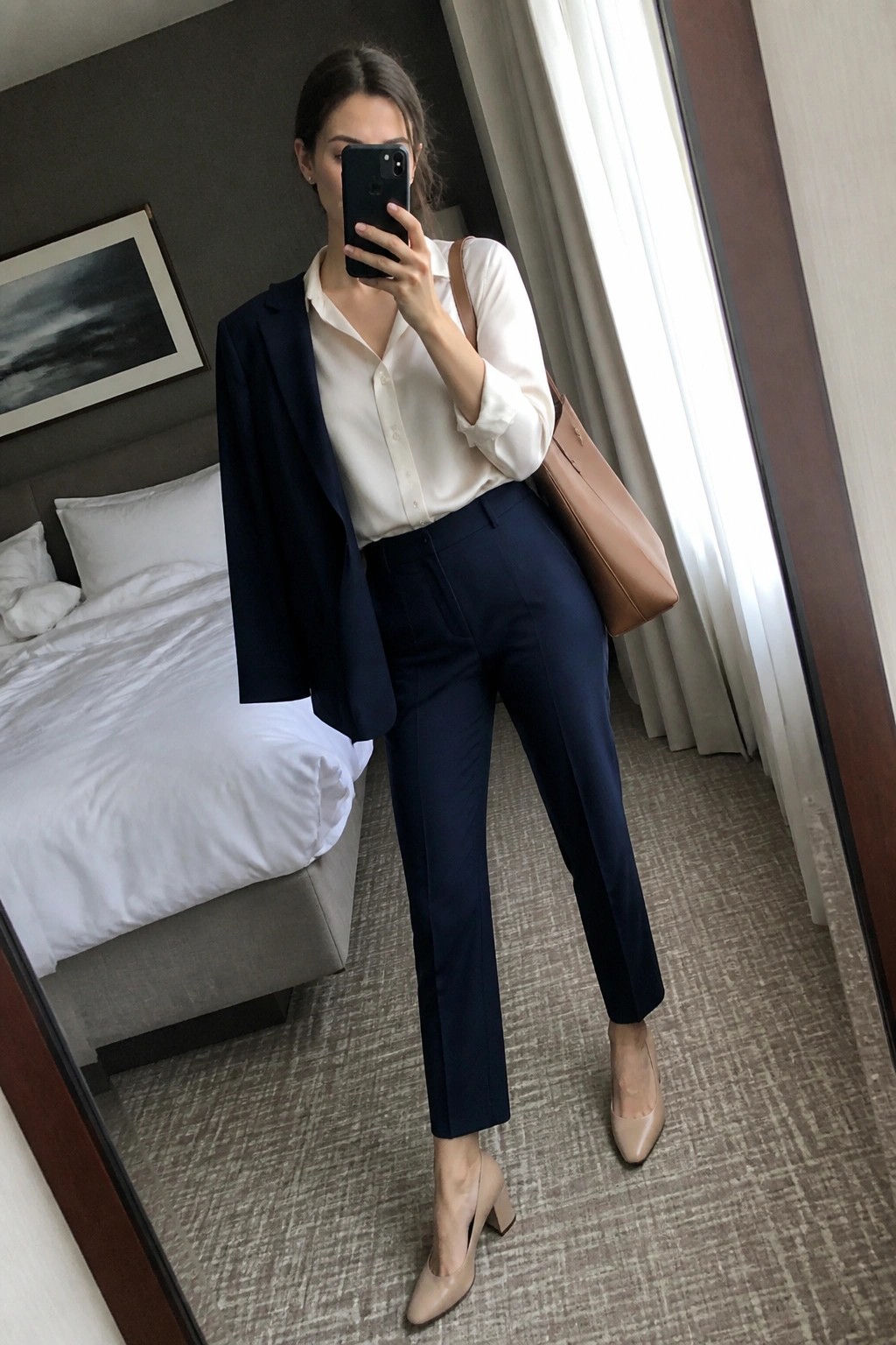 Woman wearing a navy blue tailored suit with an open blazer over a white button-up shirt, paired with navy trousers, nude block heel pumps, and a brown leather tote bag.