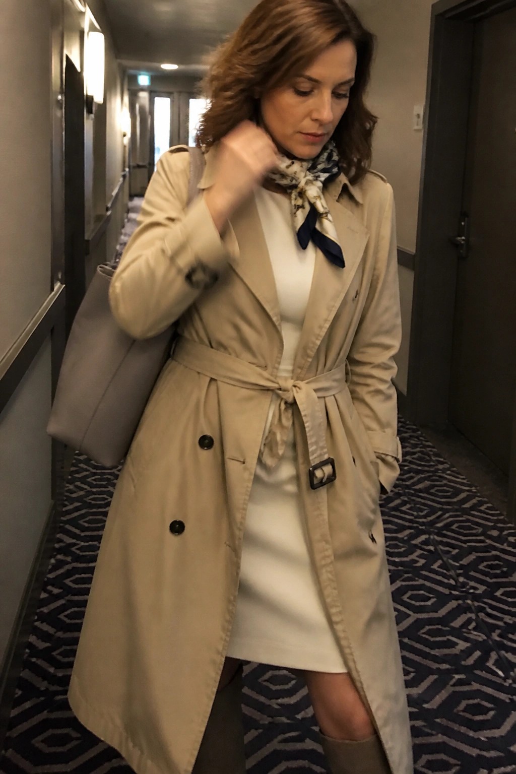 Woman in beige trench coat and scarf with gray bag in hotel hallway