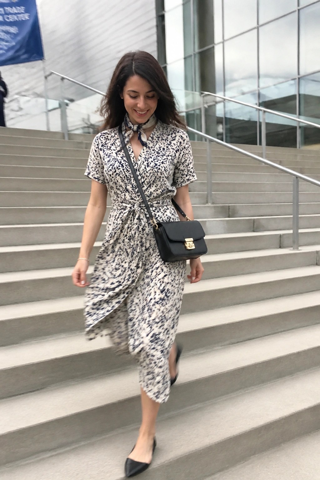 Woman wearing a short sleeve black and white abstract print wrap dress tied at the waist with a matching neck scarf, black crossbody bag, and black pointed flats while standing on concrete stairs.