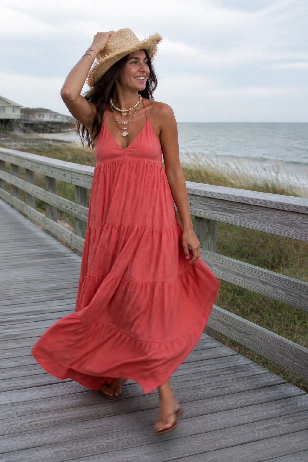 Woman wearing a coral tiered maxi dress with thin straps, layered necklaces, straw hat, and flip flops standing on a wooden boardwalk by the ocean.