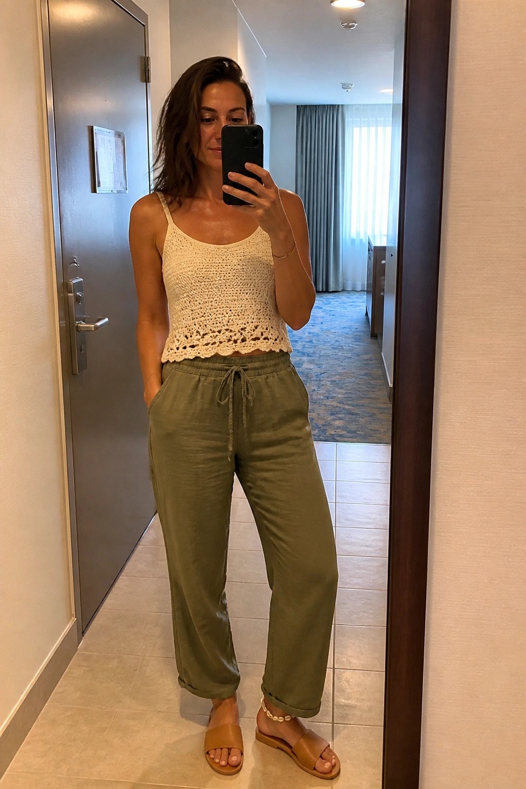Woman in white crochet top and olive pants takes mirror selfie in hallway.