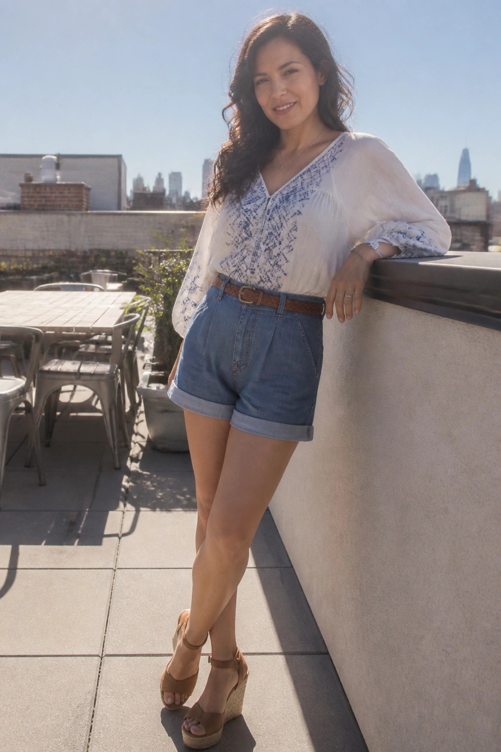 Woman in white patterned blouse and cuffed denim shorts on sunny city rooftop
