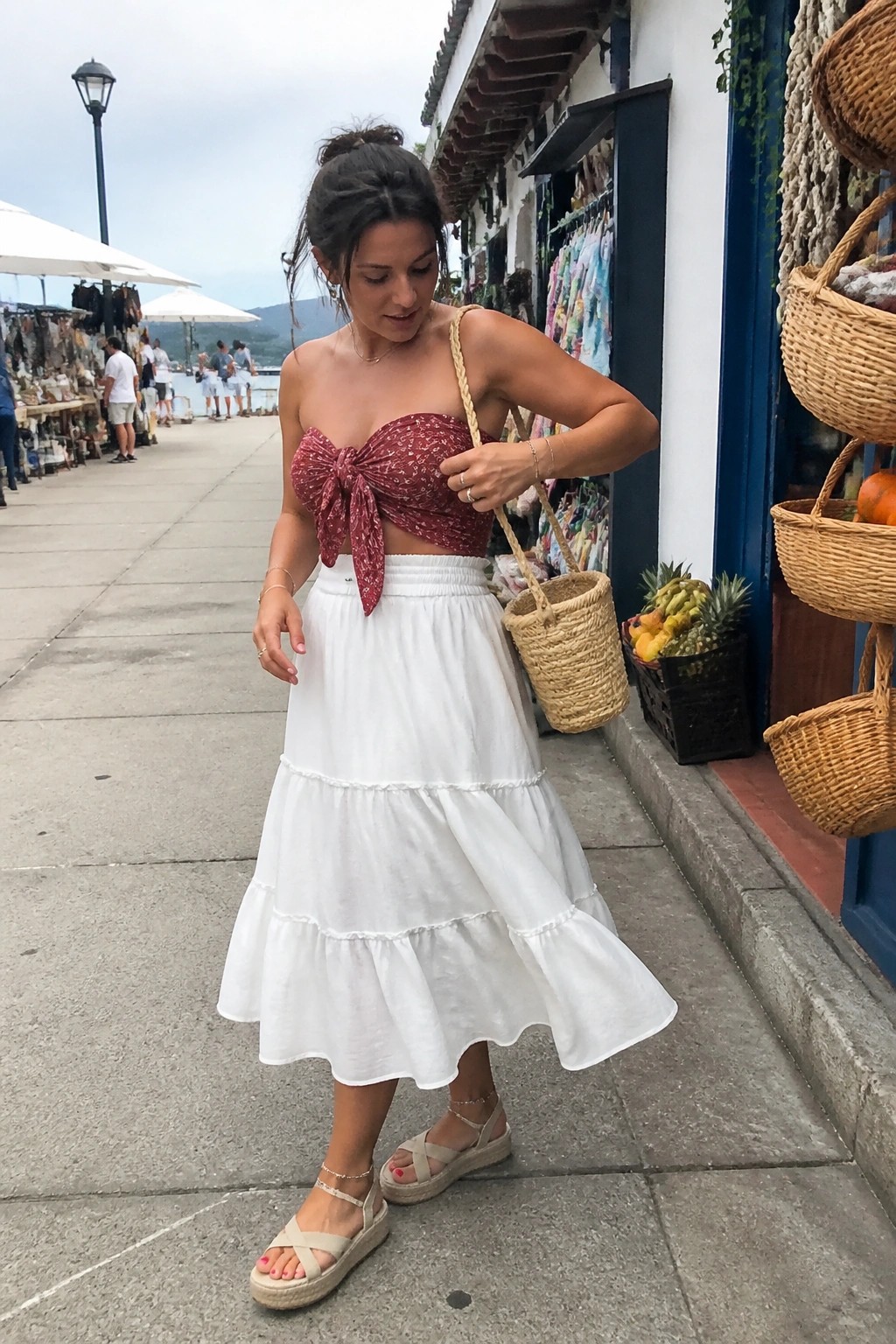 Woman in red patterned bow top and white tiered skirt holding woven bag on street