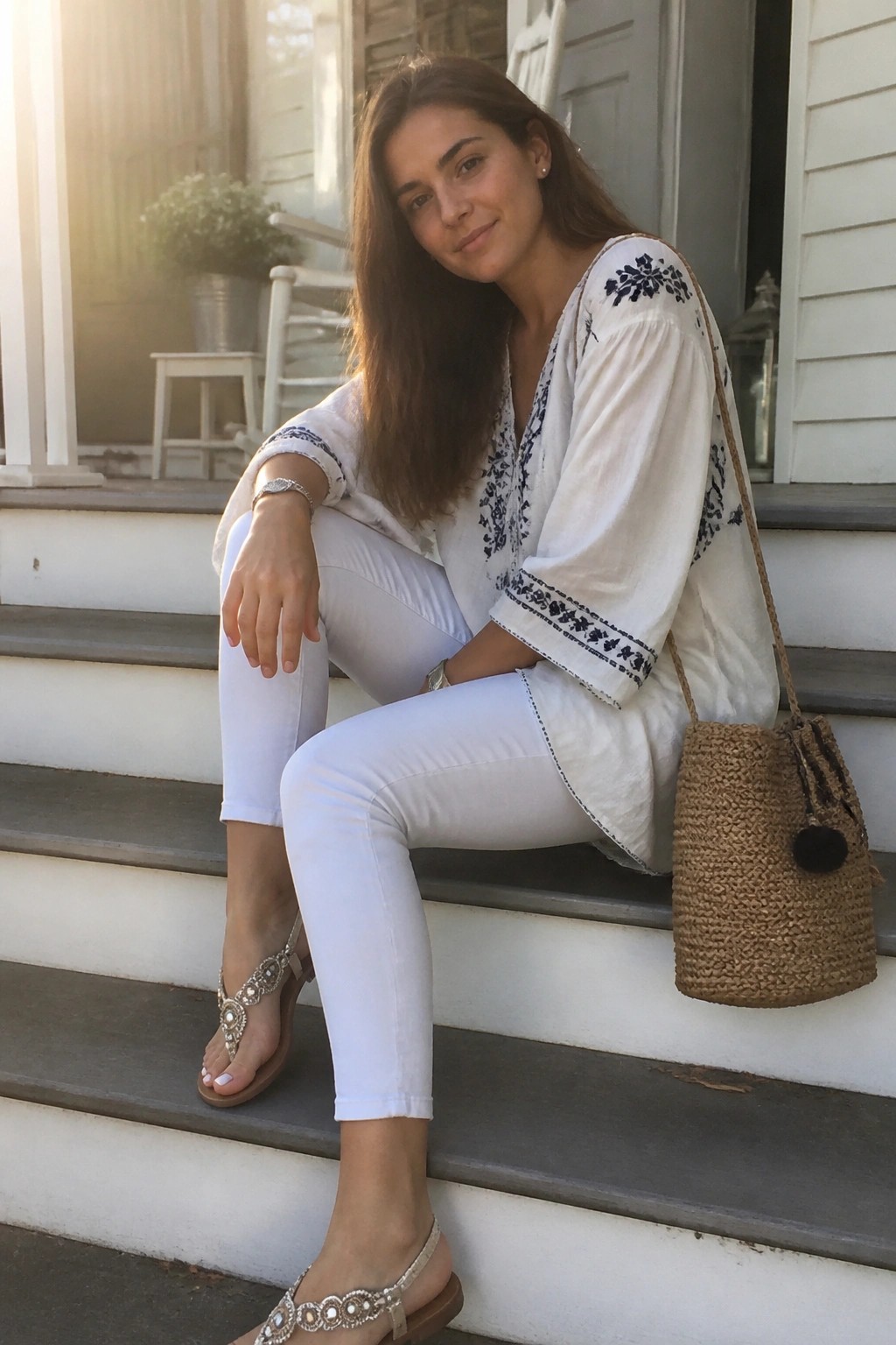 Woman in white embroidered top and pants sitting on porch steps with straw bag.