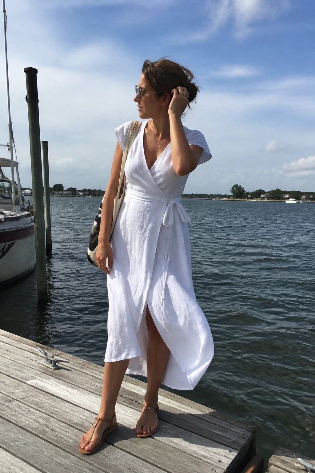 Woman in white wrap dress standing on wooden dock by calm water with boats.