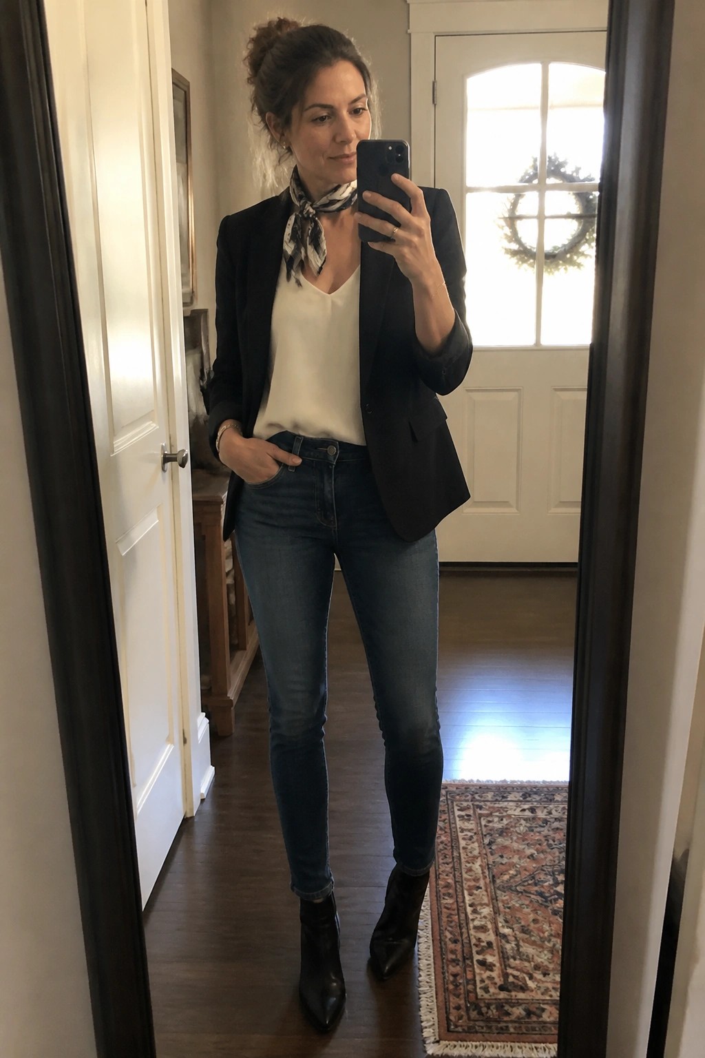 Woman in a black blazer over a white top with a patterned scarf, blue jeans, and black ankle boots taking a mirror selfie.
