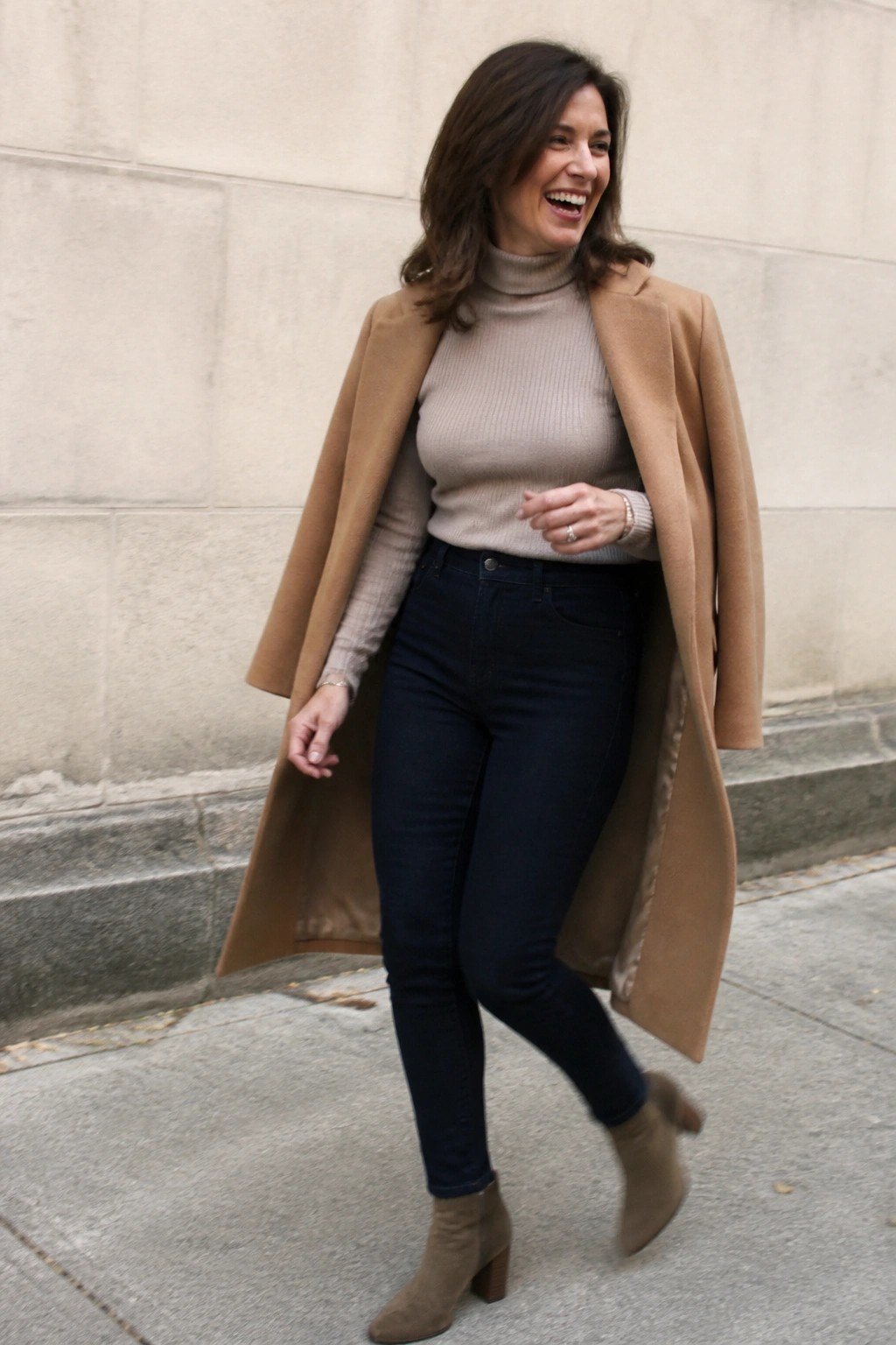 Woman in tan coat and beige turtleneck laughing on city sidewalk