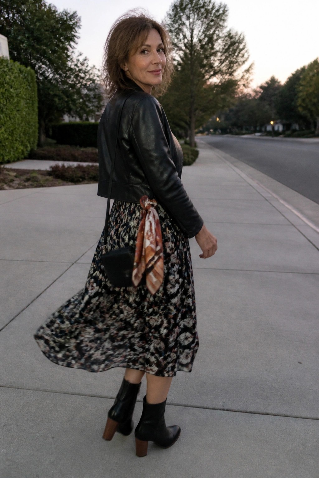Woman wearing a cropped black leather jacket over a dark floral print midi skirt with a patterned scarf tied at the waist, black ankle boots, and a small black crossbody bag.