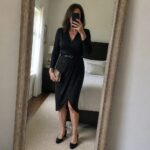 Woman in black wrap dress taking mirror selfie in bedroom holding gold clutch