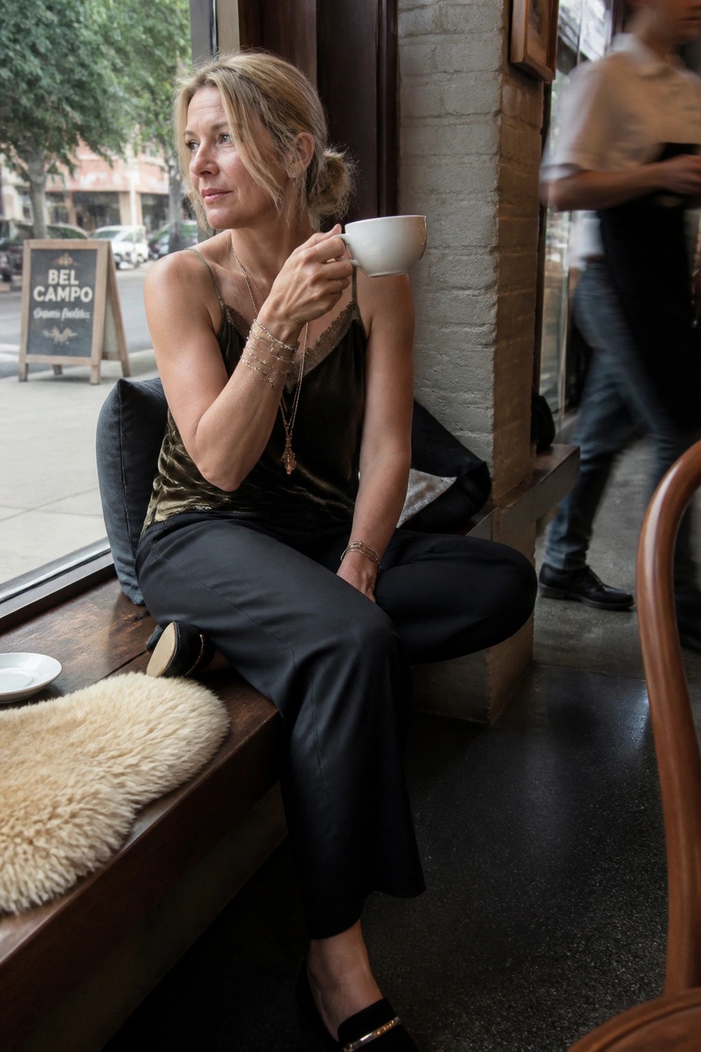 Blonde woman in green velvet top sips coffee by brick cafe window