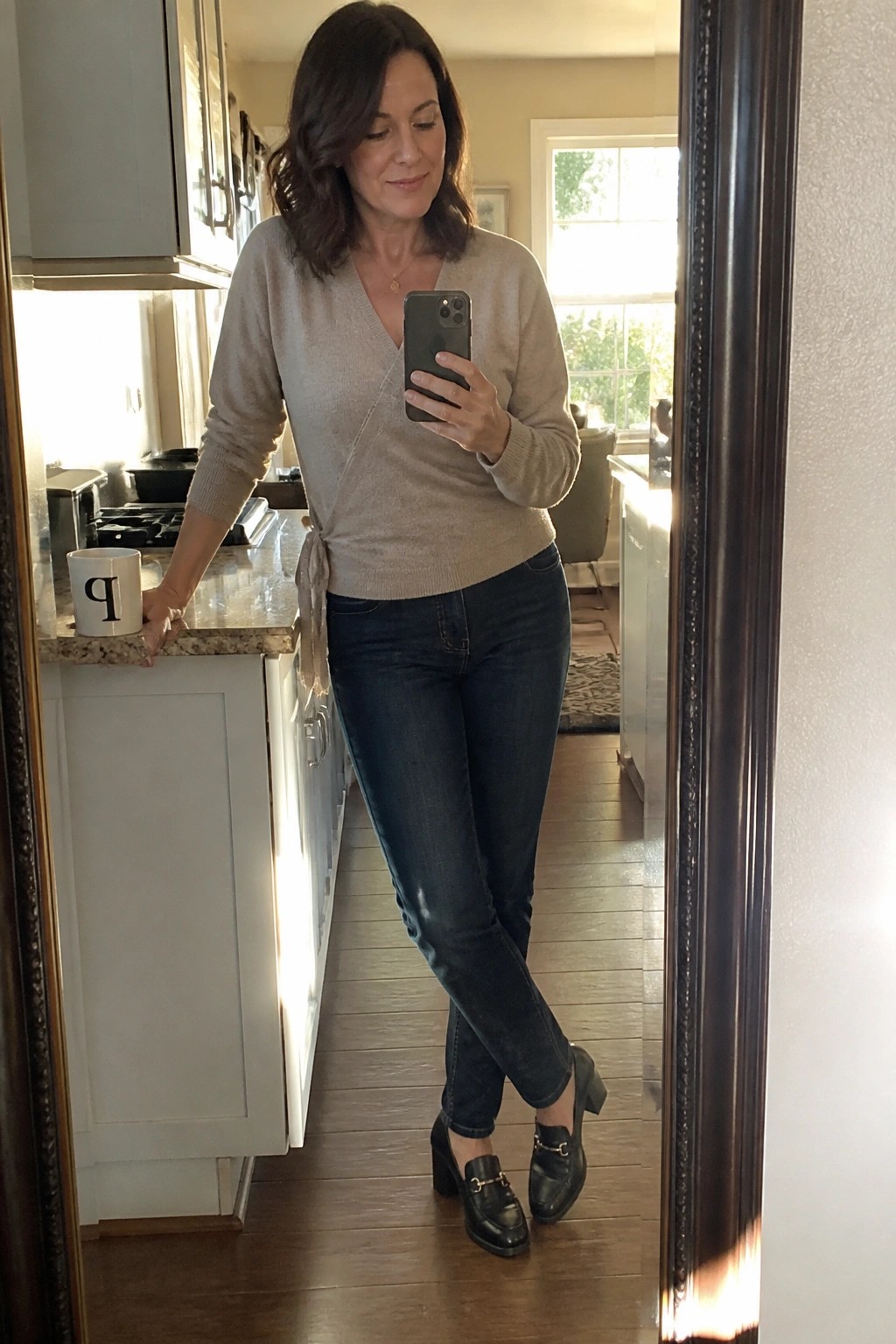 Woman in beige wrap sweater and dark jeans taking mirror selfie in kitchen