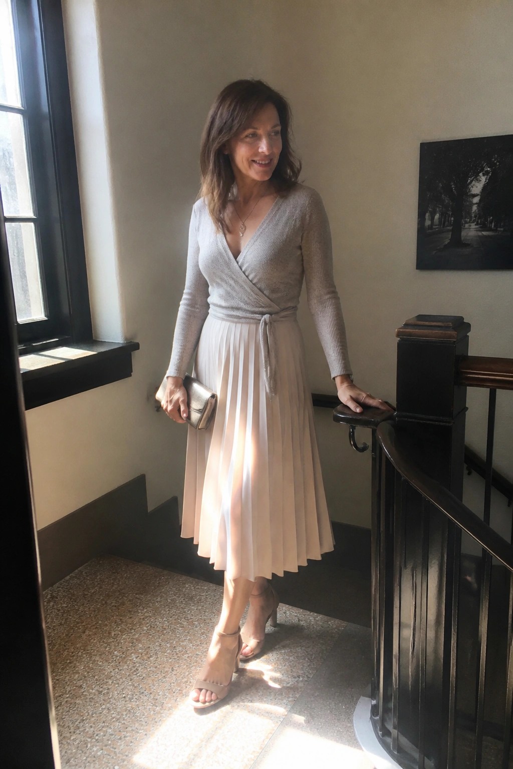 Woman in gray wrap knit sweater tied at waist over beige pleated midi skirt paired with matching heeled sandals and small metallic clutch for a date night look.