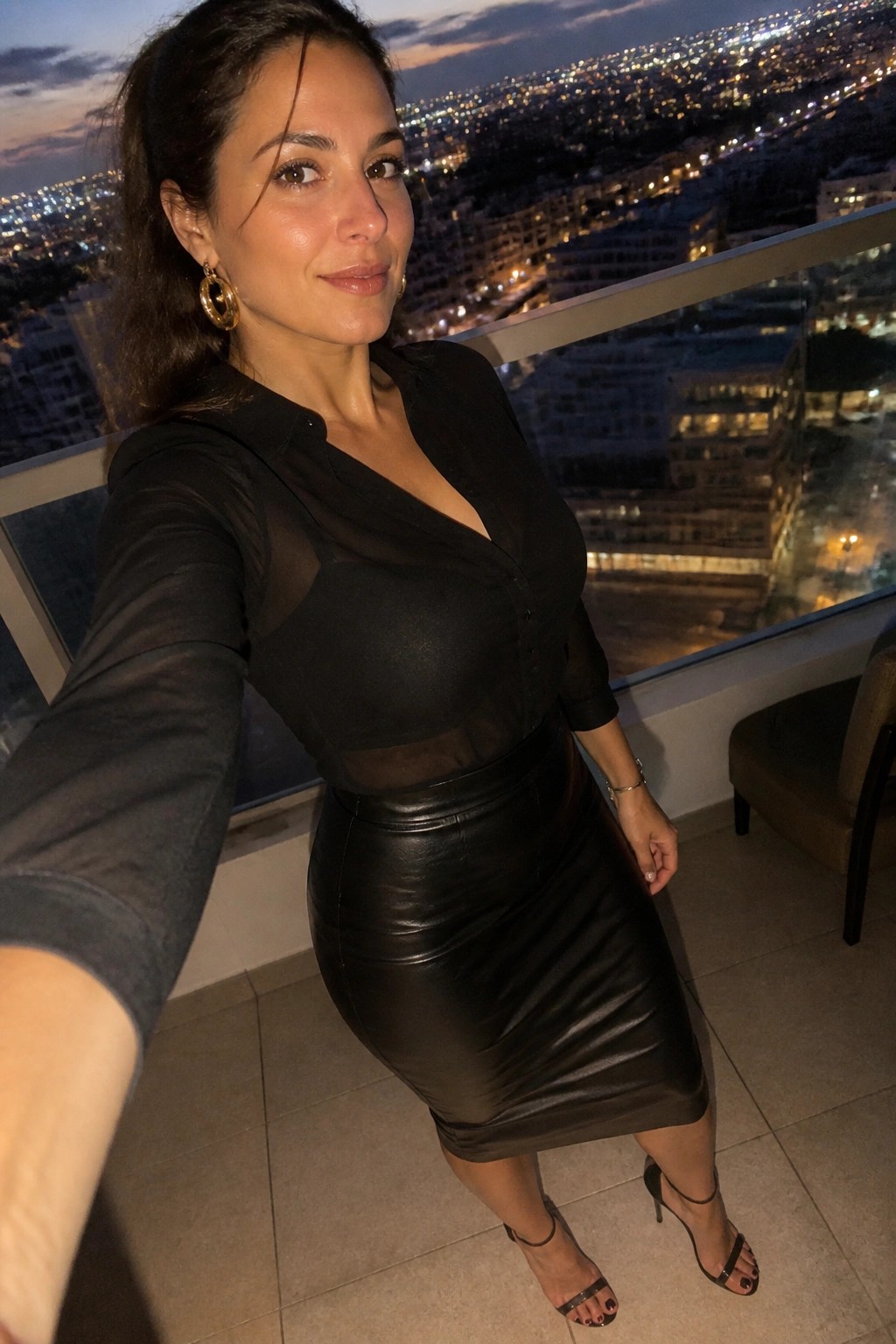 Woman in sheer black blouse and leather skirt posing on city balcony at night