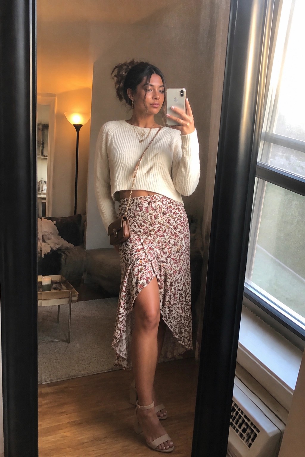 Woman taking a mirror selfie in a white cropped ribbed knit sweater paired with a floral print wrap skirt featuring an asymmetrical ruffled hem and front slit, completed with beige block heel sandals, a small brown leather crossbody bag on a gold chain strap, and layered gold necklaces.