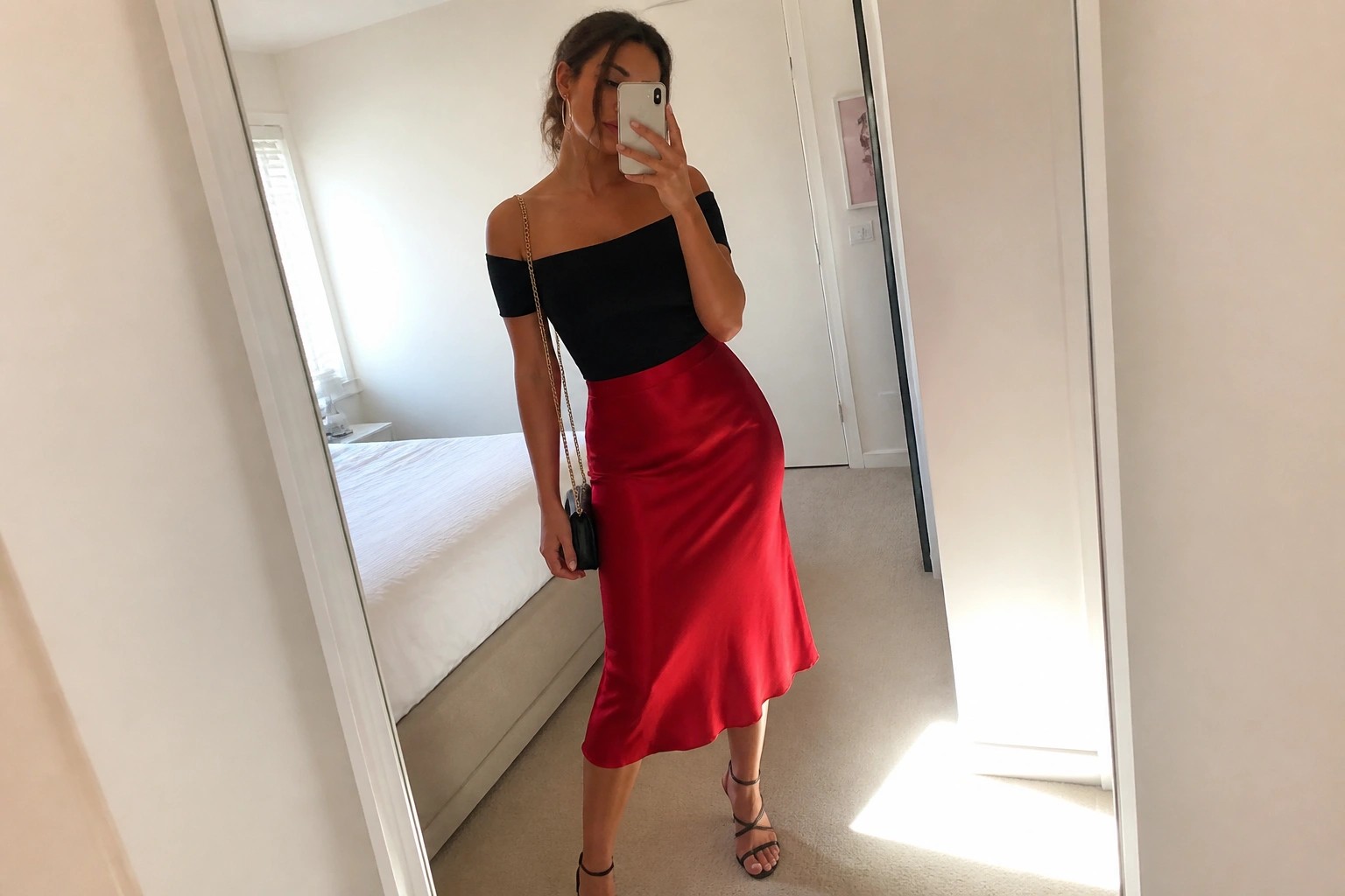 Woman in black off-shoulder top and red satin skirt taking bedroom mirror selfie