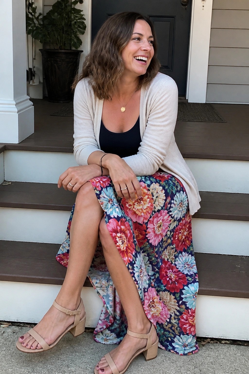 Woman wearing a long colorful floral maxi skirt with a cream cardigan over a black top and beige block heel sandals while seated on outdoor steps.