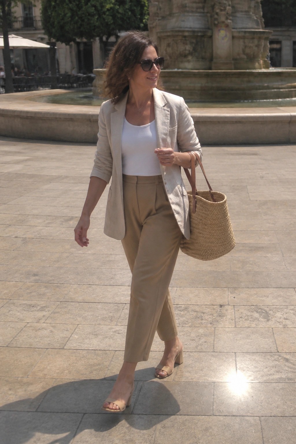Woman in beige blazer and trousers carries straw tote by sunny fountain.