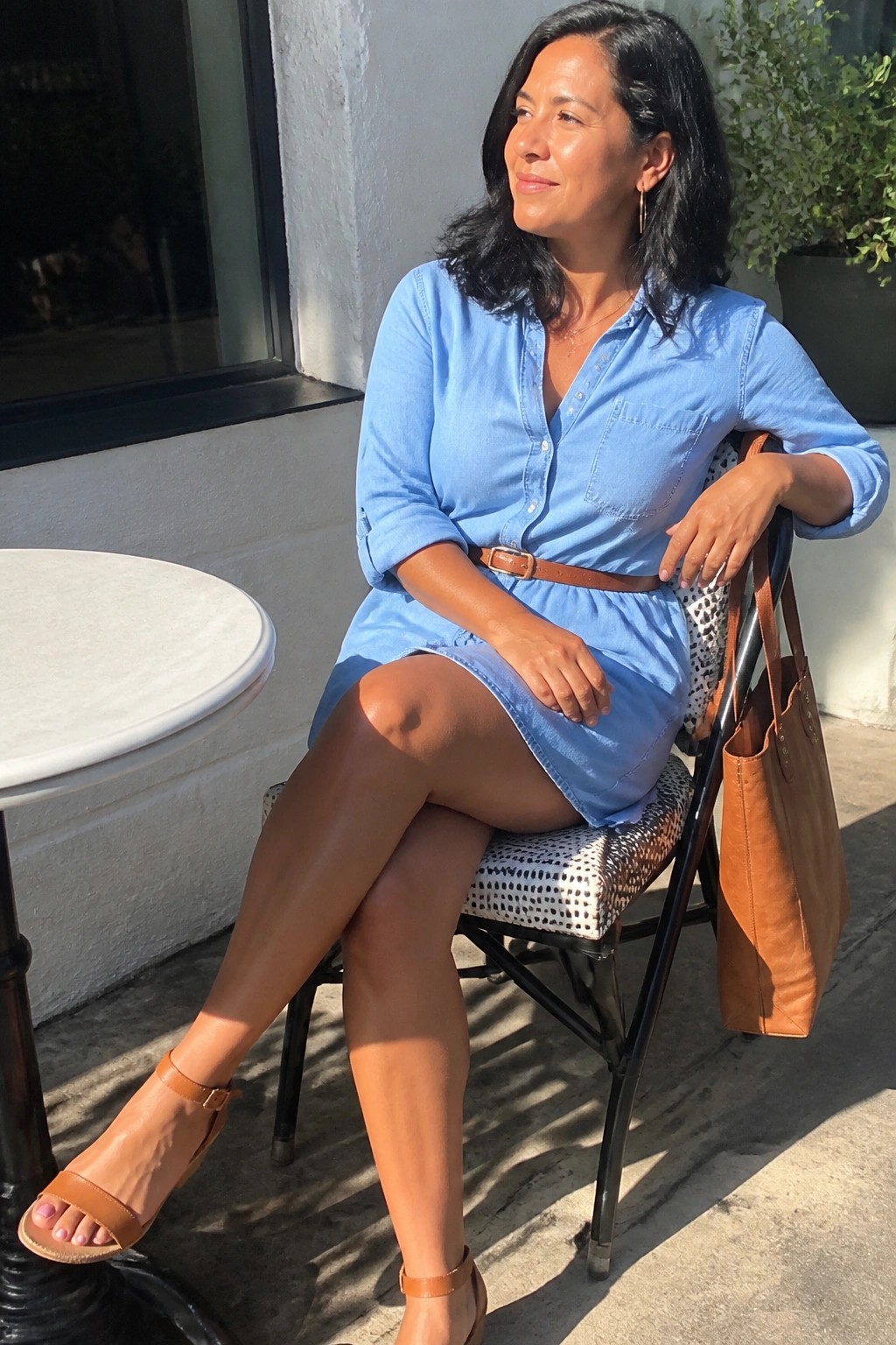 Woman in blue denim shirt dress seated outdoors with brown leather tote bag