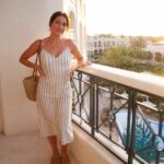 Woman in beige and white vertically striped midi dress with thin straps, tie waist, and side slit, wearing layered gold necklaces, hoop earrings, tan slide sandals, and carrying a woven straw tote bag.