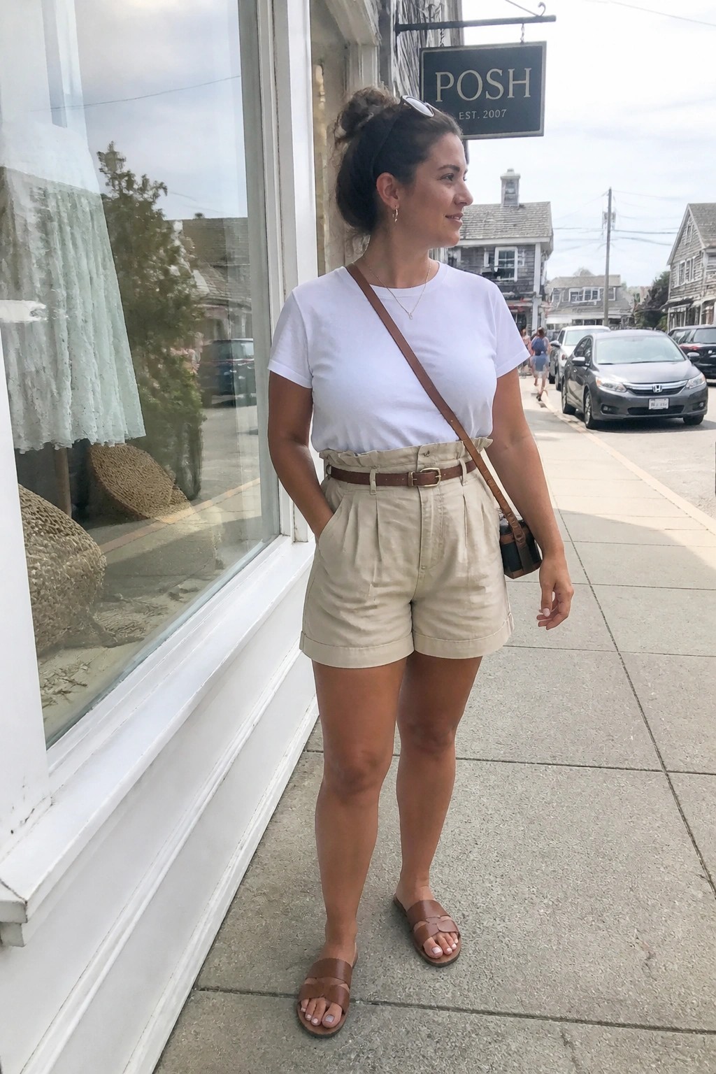 Woman in white tee and beige shorts on sunny sidewalk by storefront