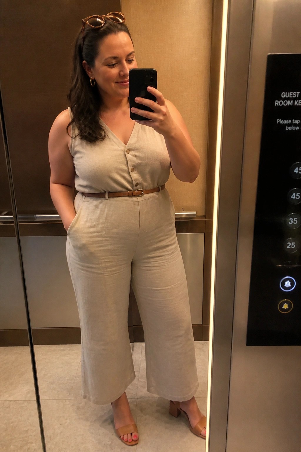 Woman wearing a beige linen sleeveless jumpsuit with V-neck button front wide cropped legs brown belt and brown block heel sandals holding a phone in an elevator mirror selfie.