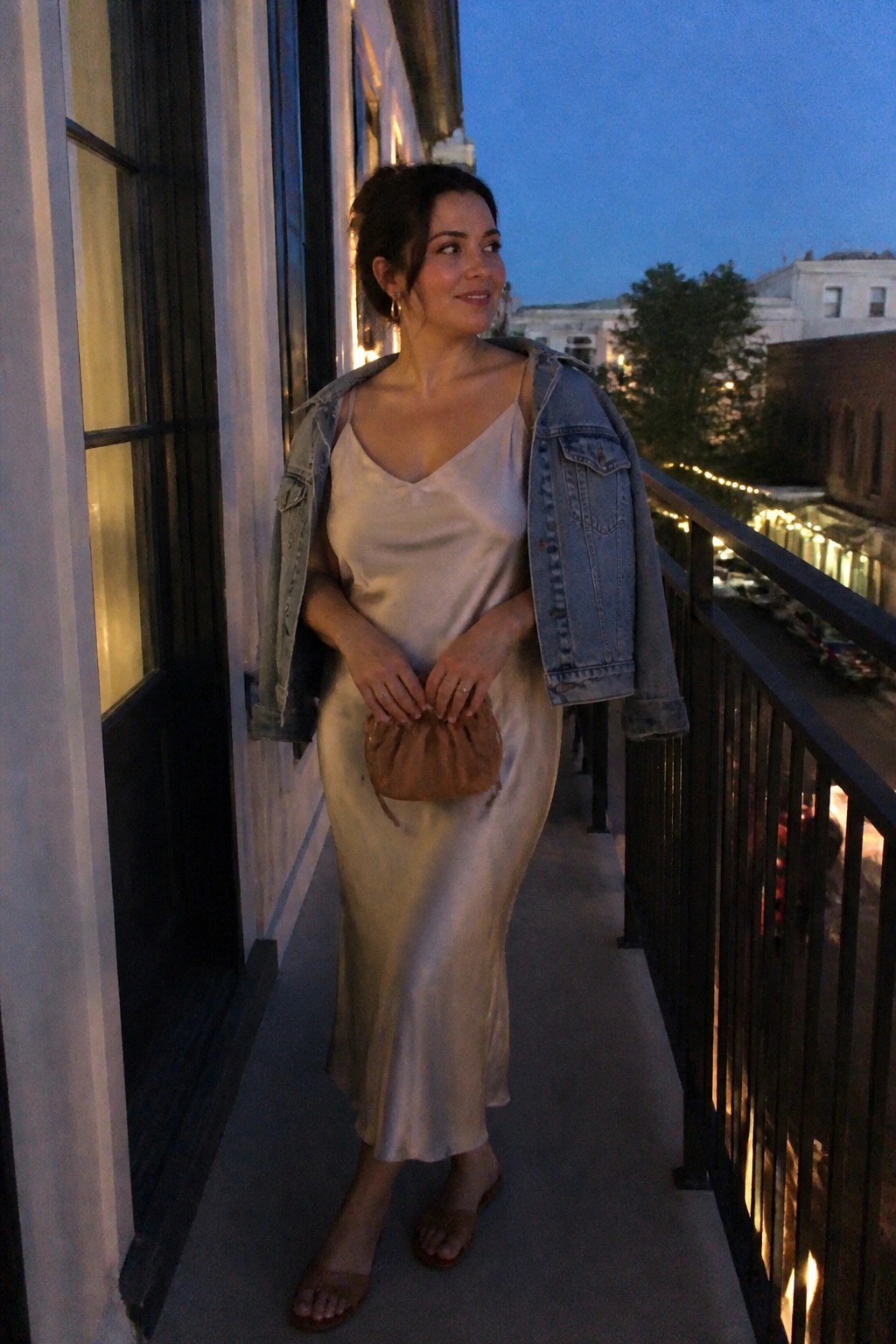 Woman in a champagne satin midi slip dress with a light wash denim jacket draped over her shoulders, brown suede clutch held in front, and matching brown slide sandals.
