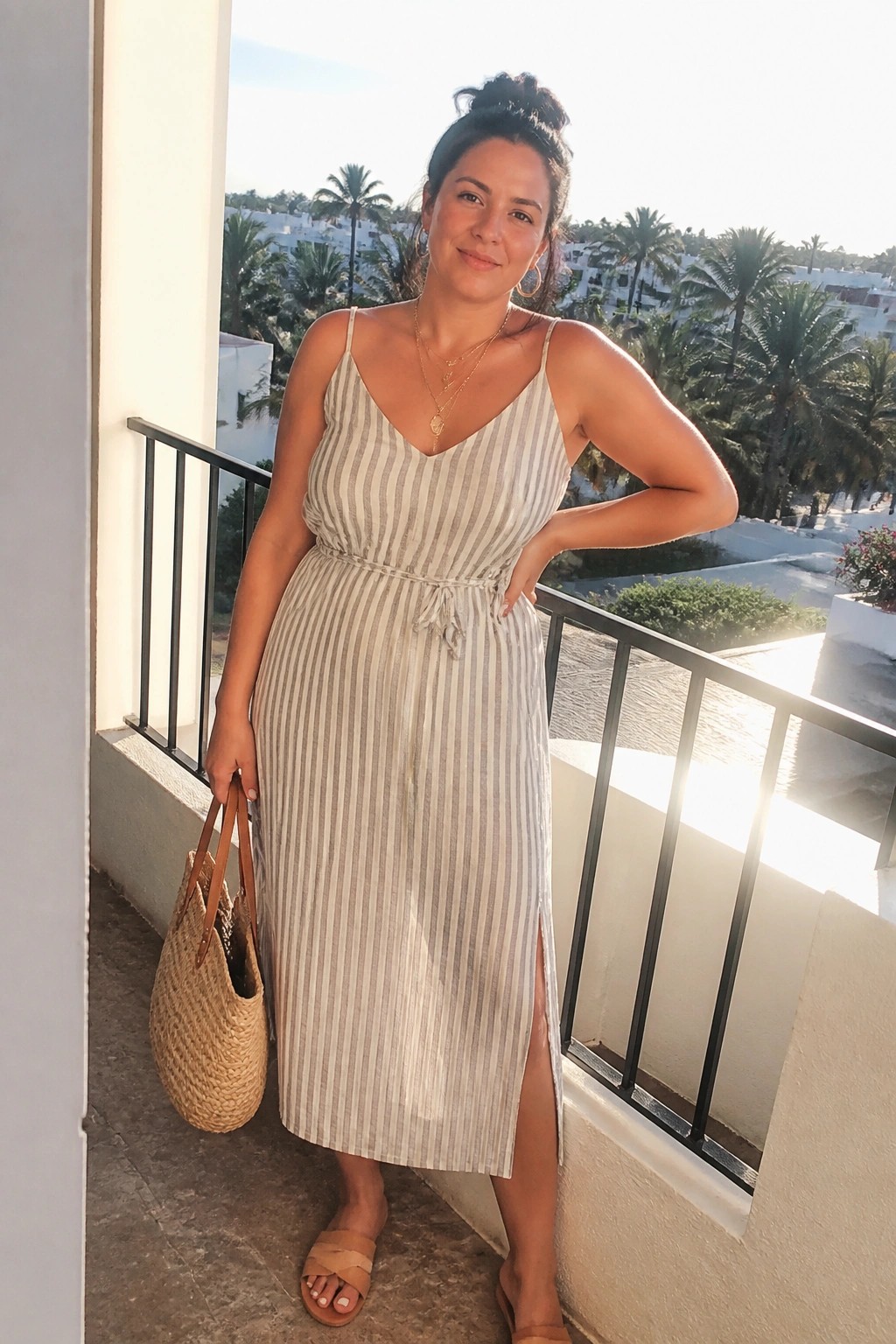 Woman in beige and white vertically striped midi dress with thin straps, tie waist, and side slit, wearing layered gold necklaces, hoop earrings, tan slide sandals, and carrying a woven straw tote bag.