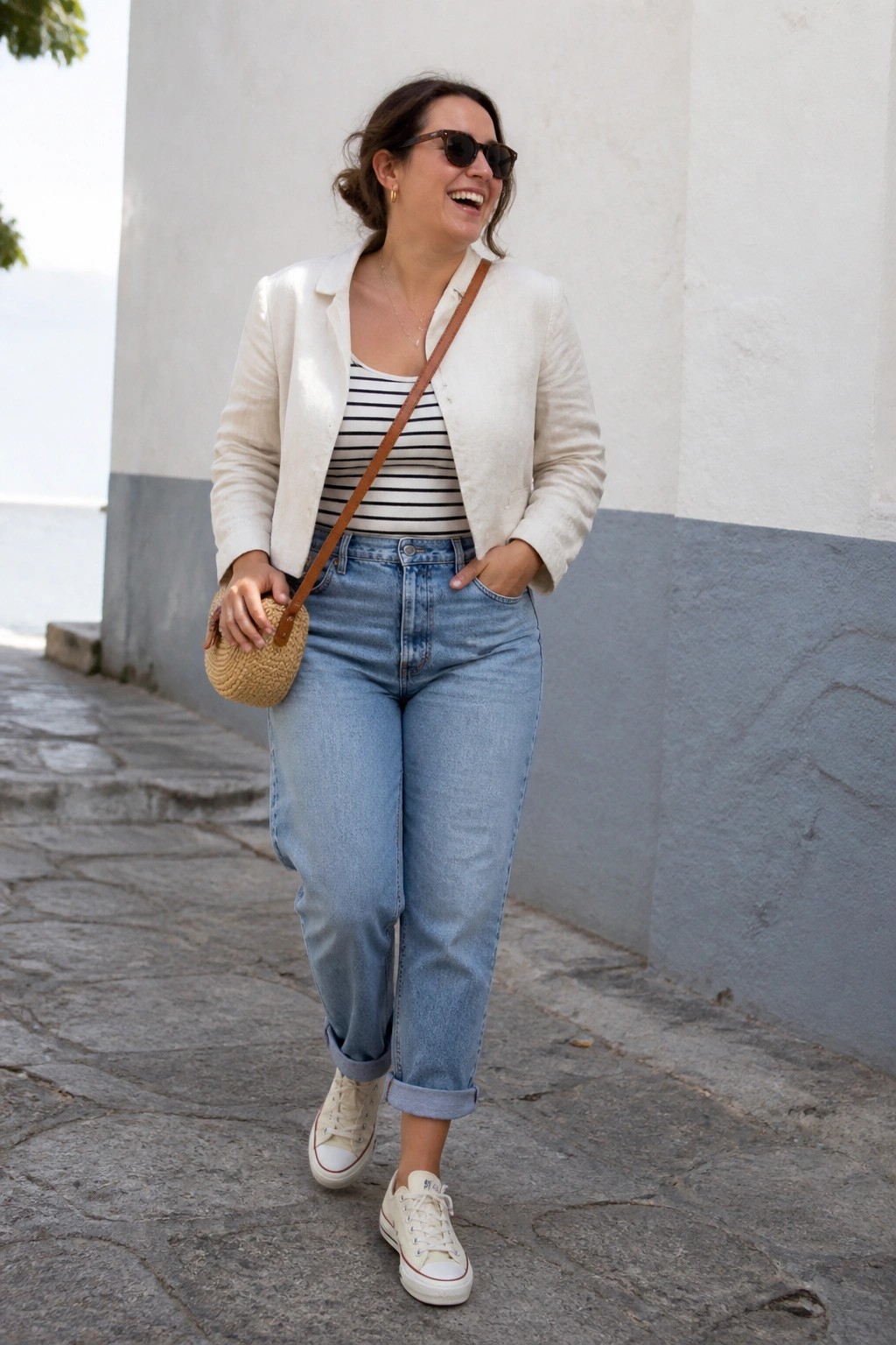 Woman wearing a white blazer over a black and white striped tank top with blue cuffed jeans and white sneakers while carrying a small woven crossbody bag.