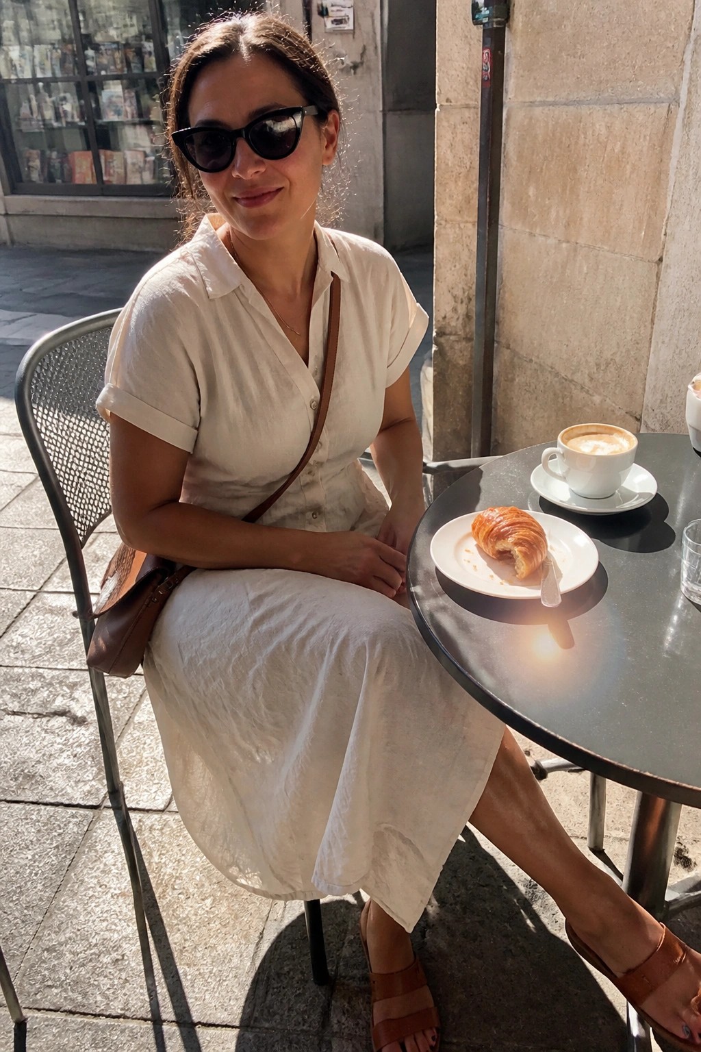 Woman in white dress and sunglasses at sunny cafe table with coffee and croissant